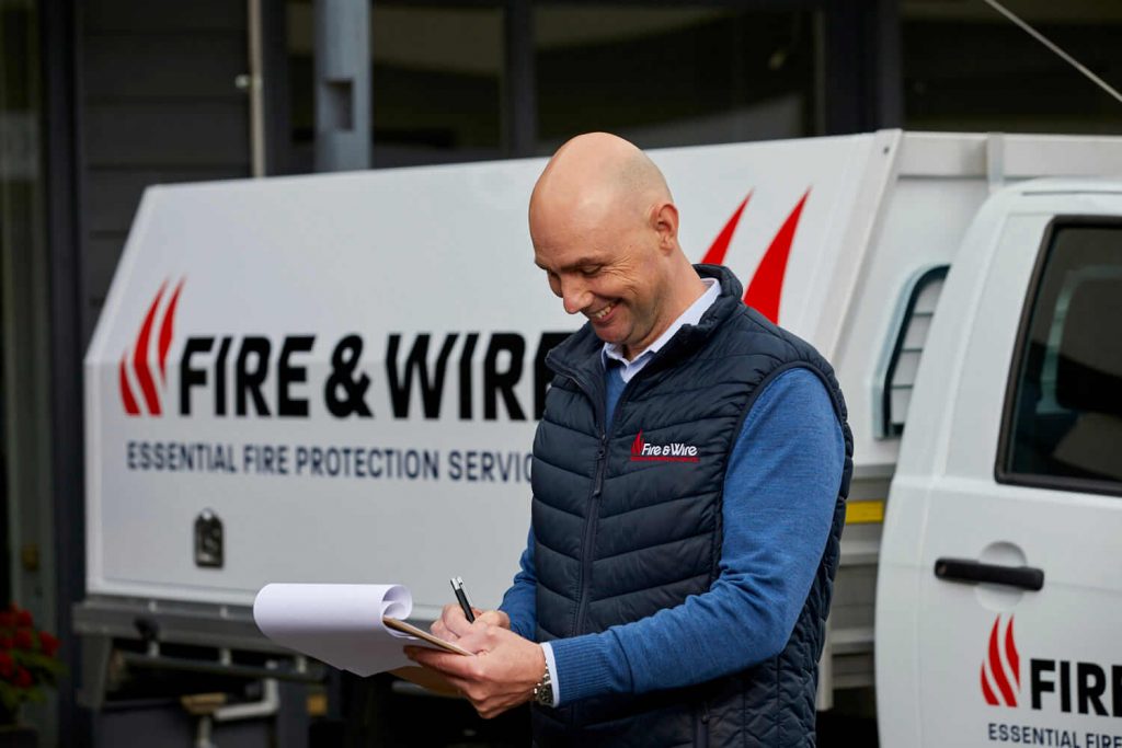 A Fire and Wire employee makes notes on a clipboard, standing in front of a Fire and Wire work vehicle