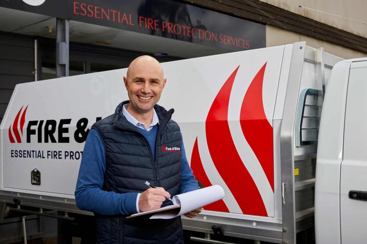 A member of the Fire and Wire fire safety training team stands in front of a company vehicle with a clipboard