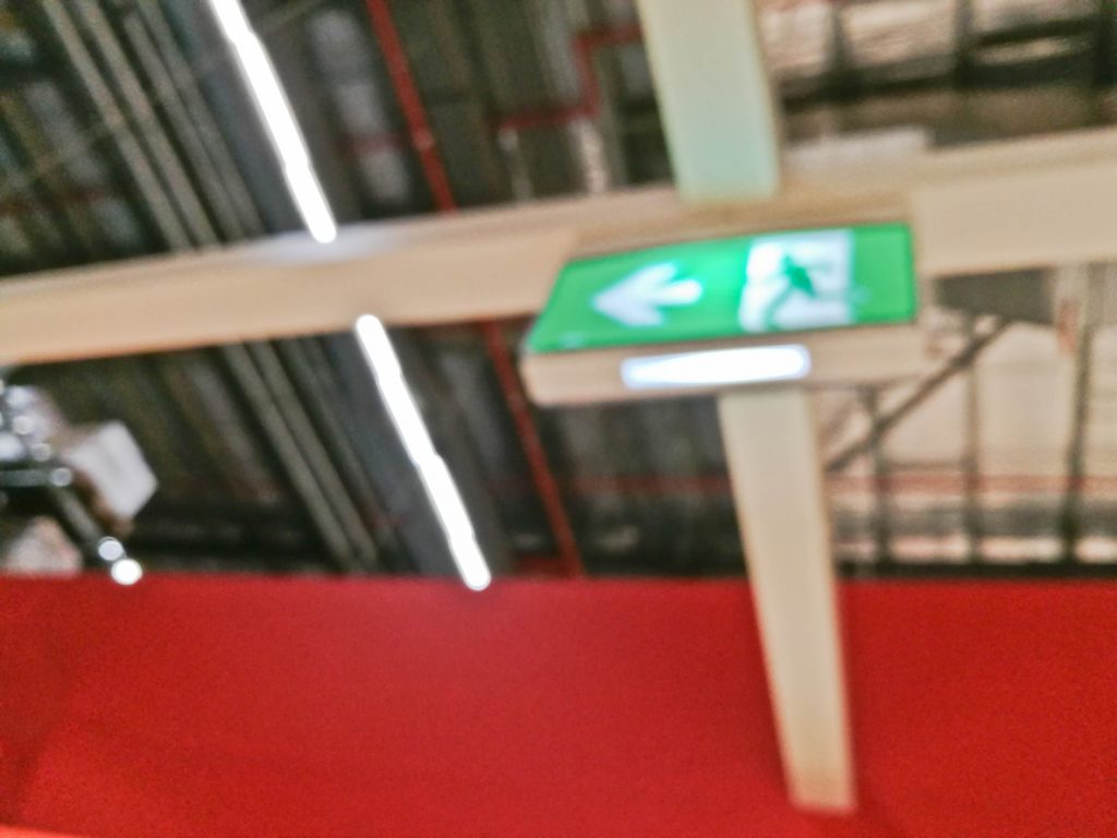 A green, illuminated emergency exit sign affixed to a warehouse ceiling